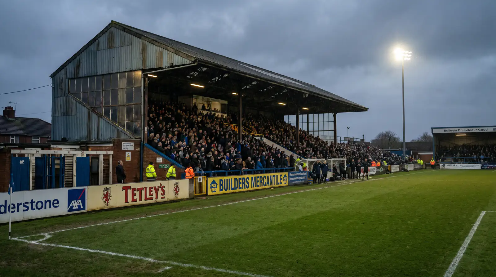 Modest League One football ground with small but passionate crowd under evening floodlights