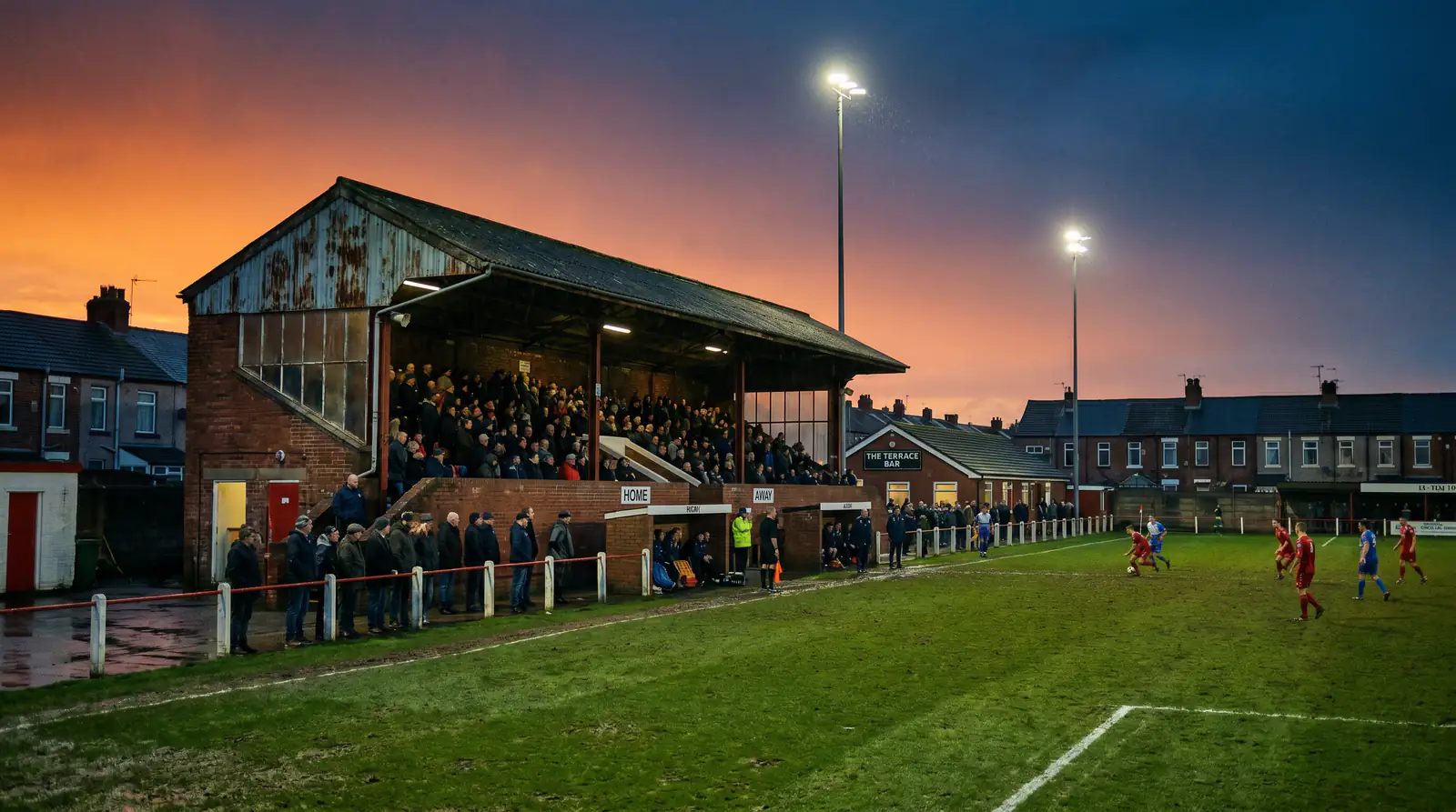 Lower-league English football ground with small crowd and floodlights during an evening match