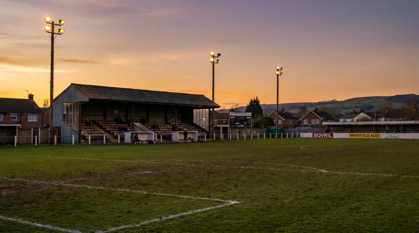Small English lower-league football ground with modest stands and floodlights at dusk